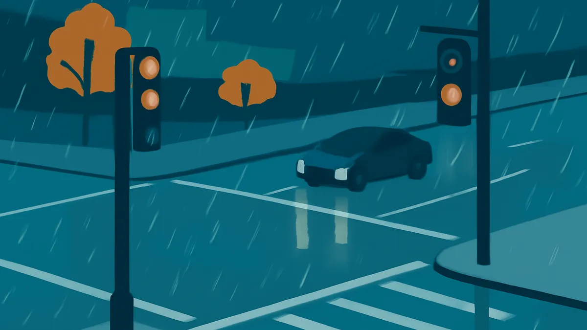 A dark blue car drives through a rainy intersection at night, with two traffic lights showing orange