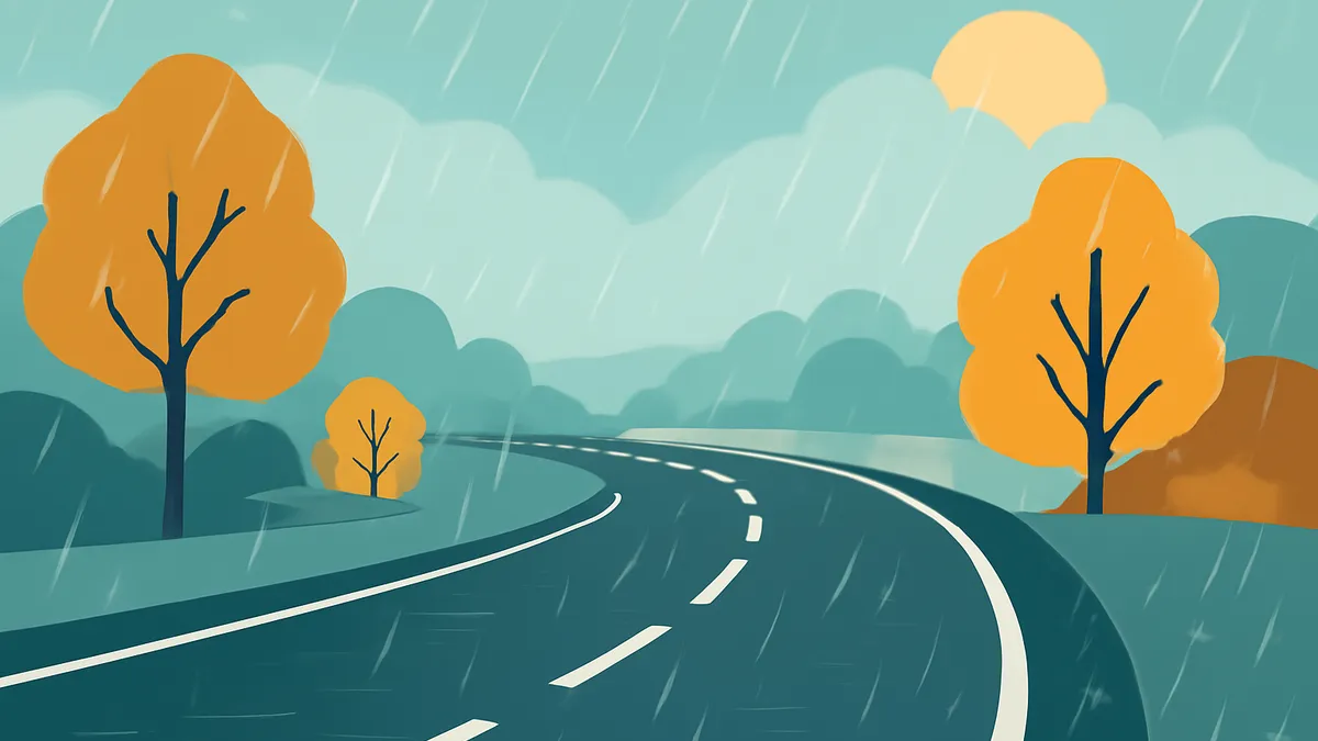 A winding road with white dashed lines on a rainy day, flanked by trees with orange foliage and green hills under a cloudy